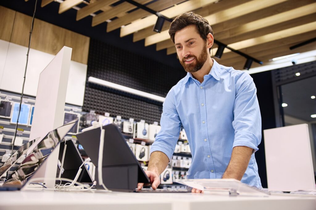A man browsing laptops and tablets in-store, exploring the best tech deals 2025, tablet discounts 2025, AirPods sale 2025, top gadget deals, best accessories for laptops, and tech sales for students in a sleek electronics retail environment.