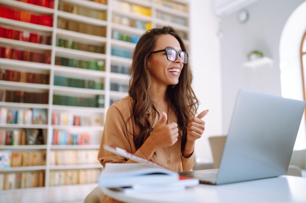 Smiling professional woman giving a thumbs up while working on a laptop in a bright office library, representing a positive comparison of Dell XPS vs MacBook in 2025 for corporate users.