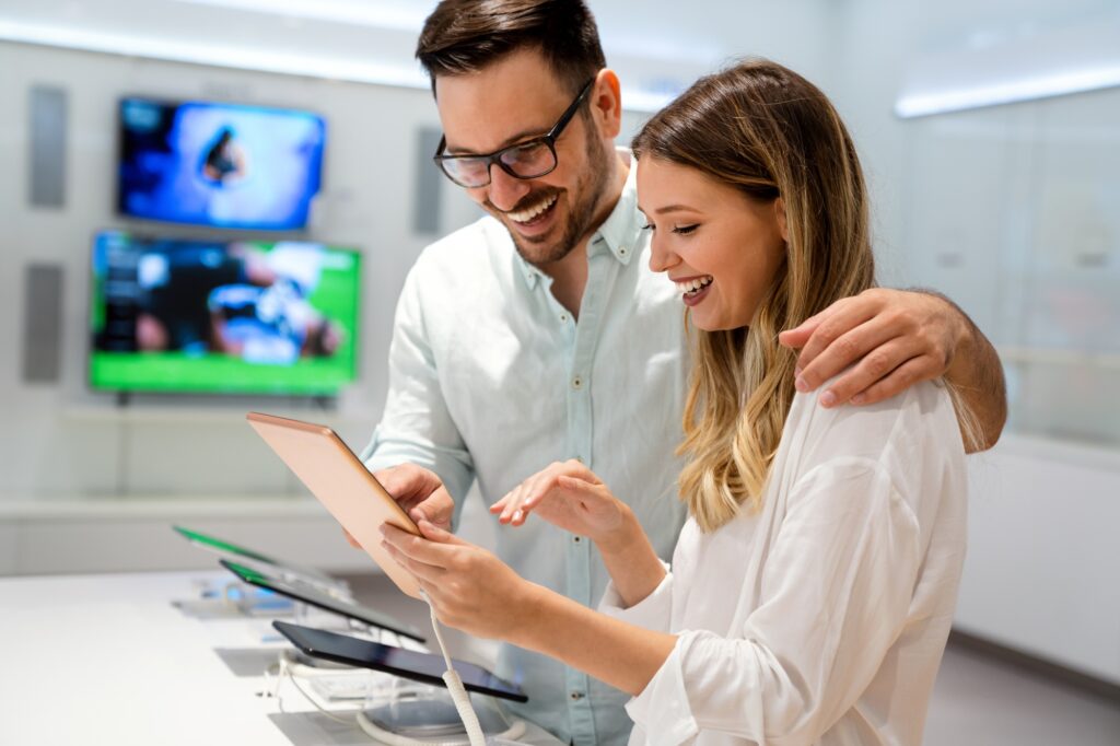 Two happy customers smiling while browsing the best tech deals 2025 on a tablet, exploring tablet discounts 2025, AirPods sale 2025, top gadget deals, best accessories for laptops, and tech sales for students in a bright, modern electronics store.