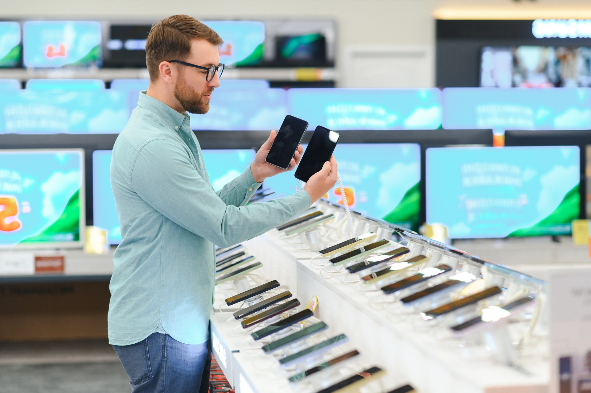 A customer comparing smartphones in an electronics store with TV displays in the background — ideal support visual for Contact UnitedTech Deals page highlighting tech product inquiries and customer service engagement.

