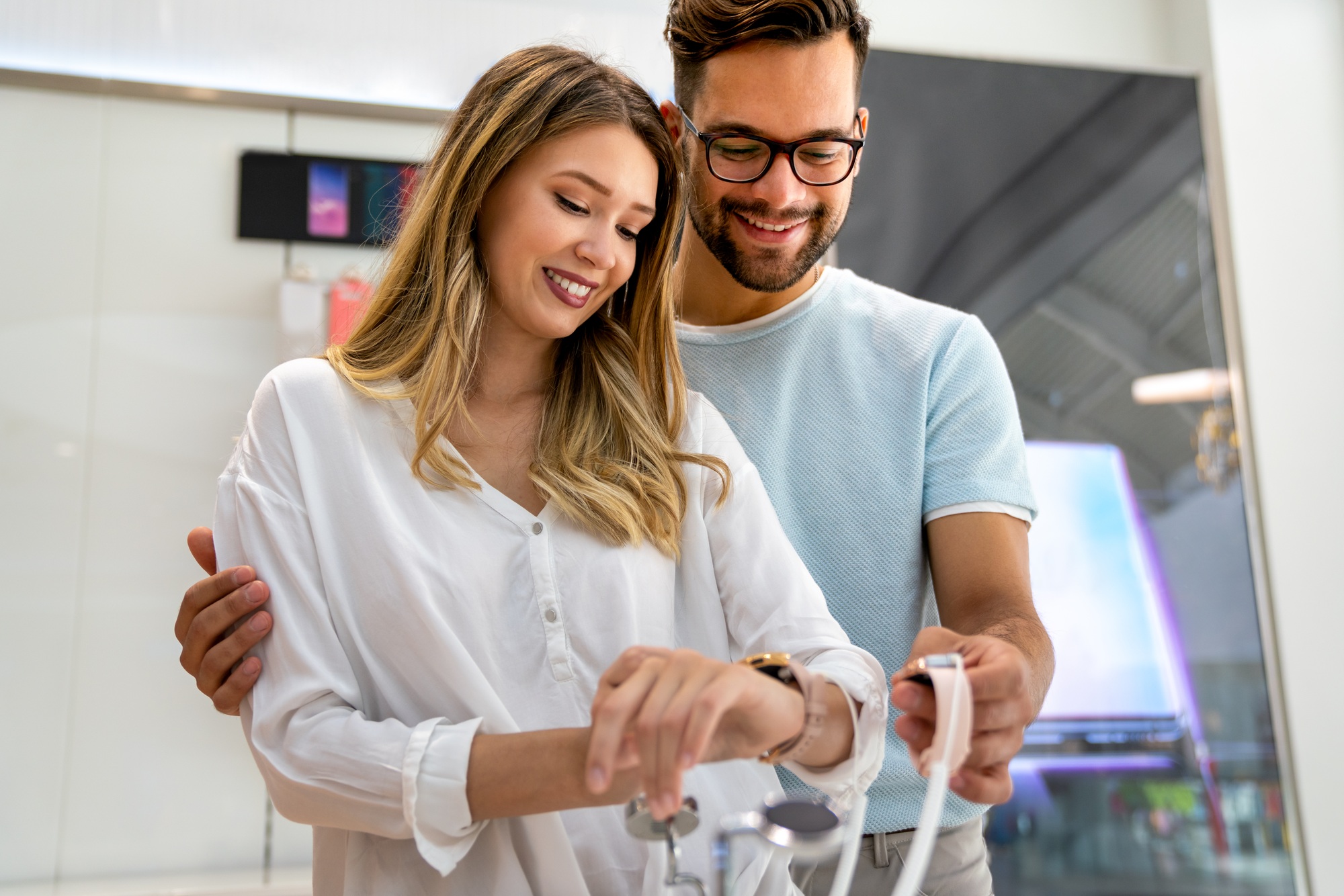 Smiling couple trying on smartwatches during a tech sale in Florida, enjoying hands-on deals at a modern electronics store. 